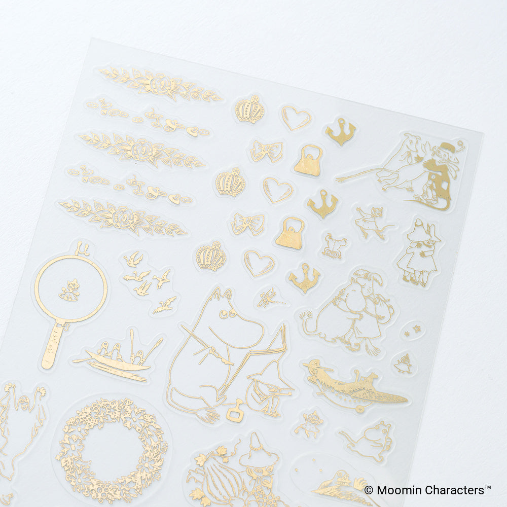 A sheet of transparent stickers featuring gold foil designs including Moomin characters, crowns, hearts, bows, and floral wreaths, displayed on a clean white background.