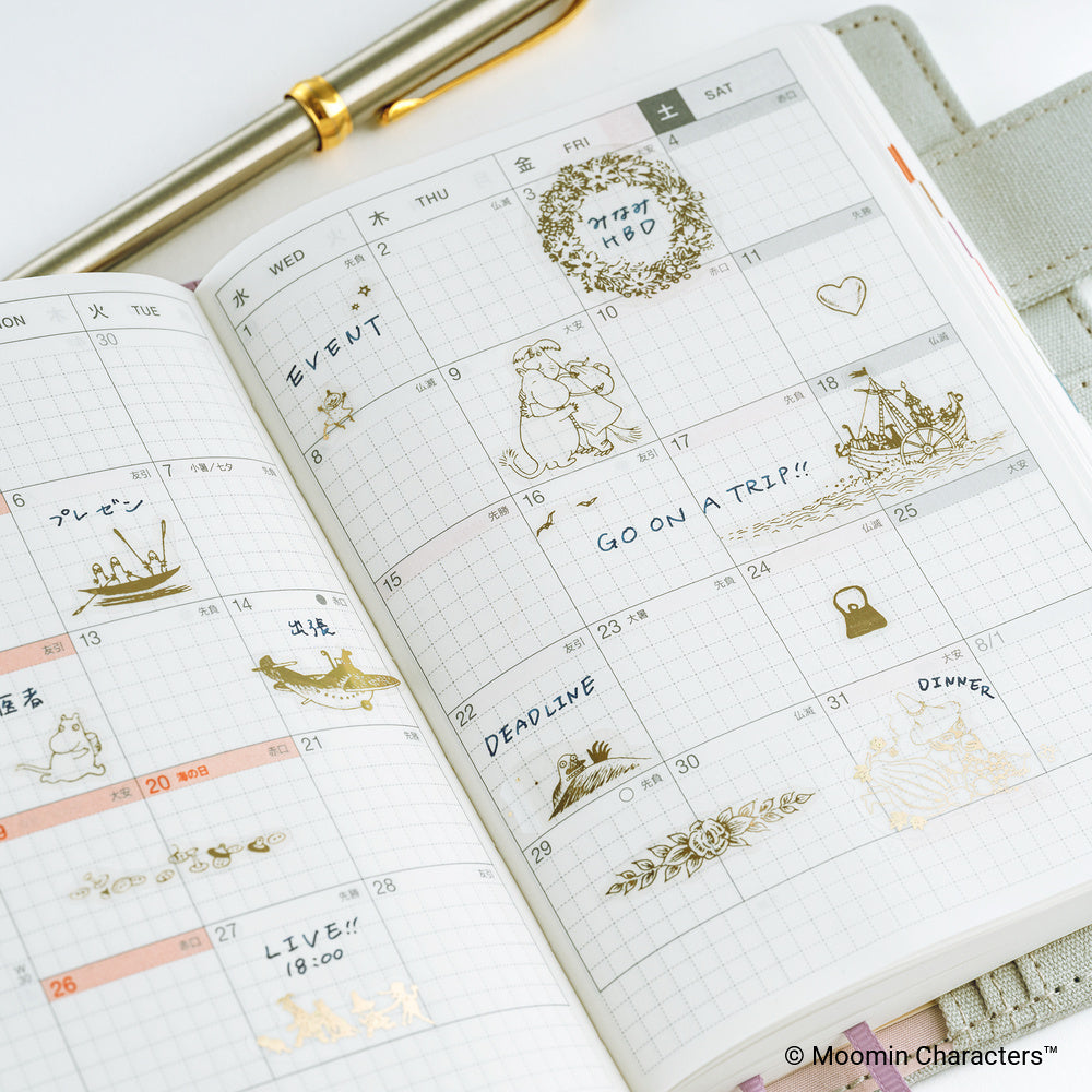 An open grid planner showing a decorated weekly calendar spread with gold foil Moomin stickers and handwritten notes, photographed on a light surface with part of a gold pen visible.