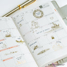 An open grid planner showing a decorated weekly calendar spread with gold foil Moomin stickers and handwritten notes, photographed on a light surface with part of a gold pen visible.