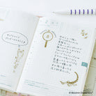An open planner with Japanese text, grid pages, decorative doodles, gold stickers, and a purple-and-white striped pen on a light surface.