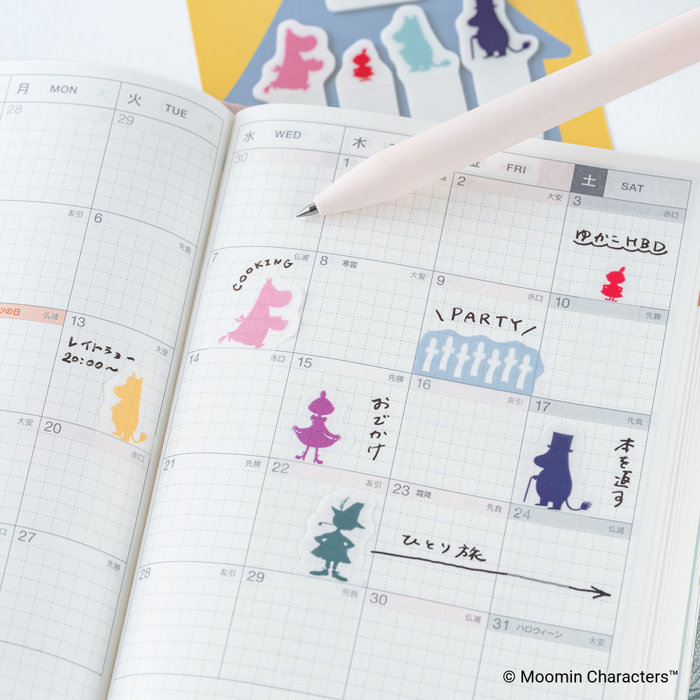 An open planner showing a weekly calendar layout with colorful Moomin character stickers placed on the pages, alongside a pale pink pen. 