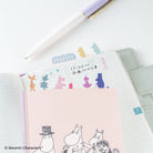 A pink Moomin Character notebook with black-and-white line art of characters and colorful silhouette index tabs, placed on top of an open grid planner. A white pen with a lavender and gold cap rests diagonally across the scene.
