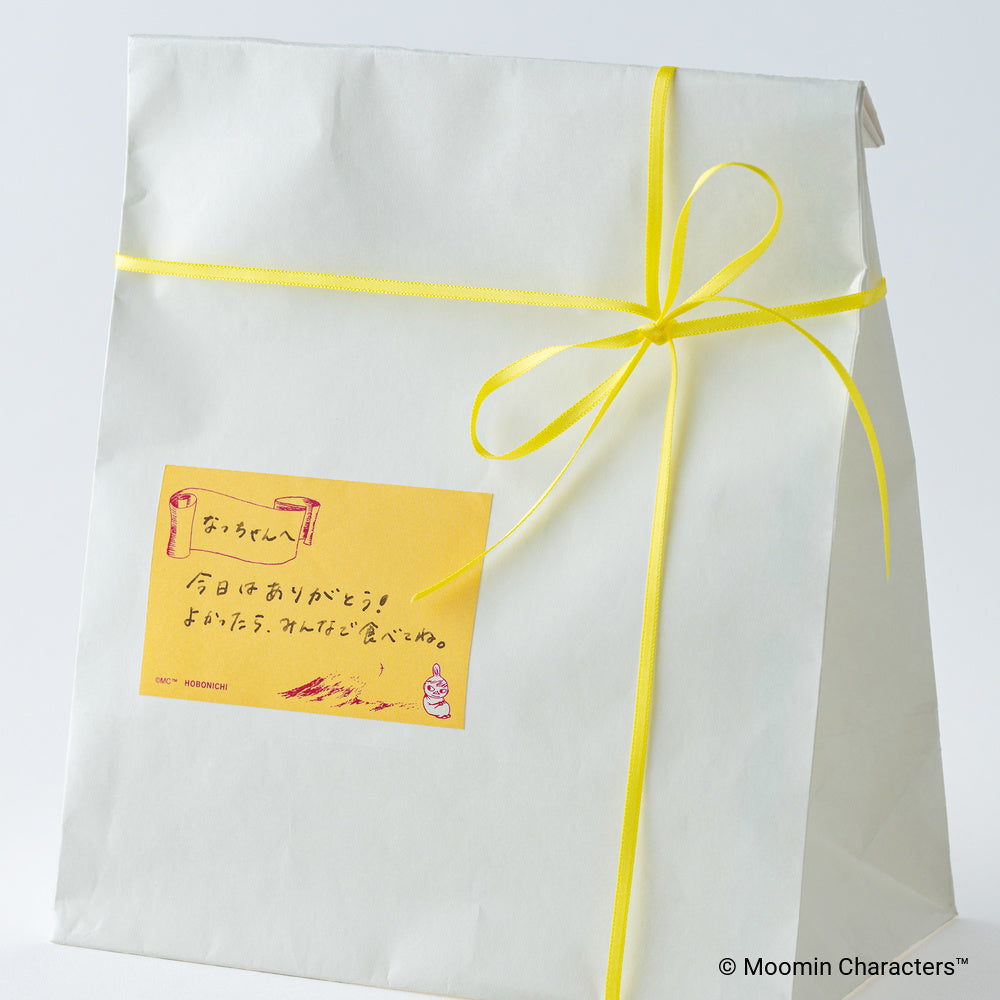 A white paper gift bag tied with a bright yellow ribbon and bow, featuring a small yellow sticky note with Japanese text and a Moomin cartoon character.