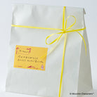 A white paper gift bag tied with a bright yellow ribbon and bow, featuring a small yellow sticky note with Japanese text and a Moomin cartoon character.