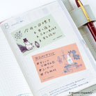 An open planner or notebook page with Japanese text, pastel sticky notes, and Moomin-themed illustrations, styled with a pen on a light fabric surface. 