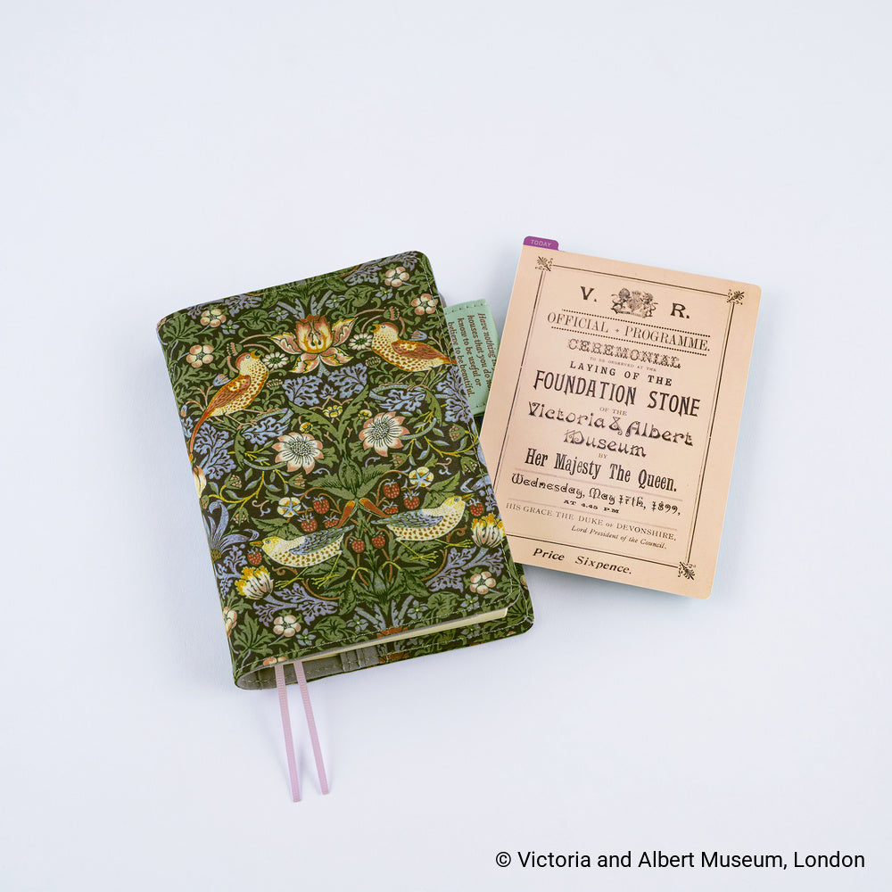 An ornate green fabric-covered notebook with a floral and bird pattern, shown next to a pencil board reading 'Official Programme' for the Victoria & Albert Museum. The notebook has lilac bookmarks, photographed on a clean white background.