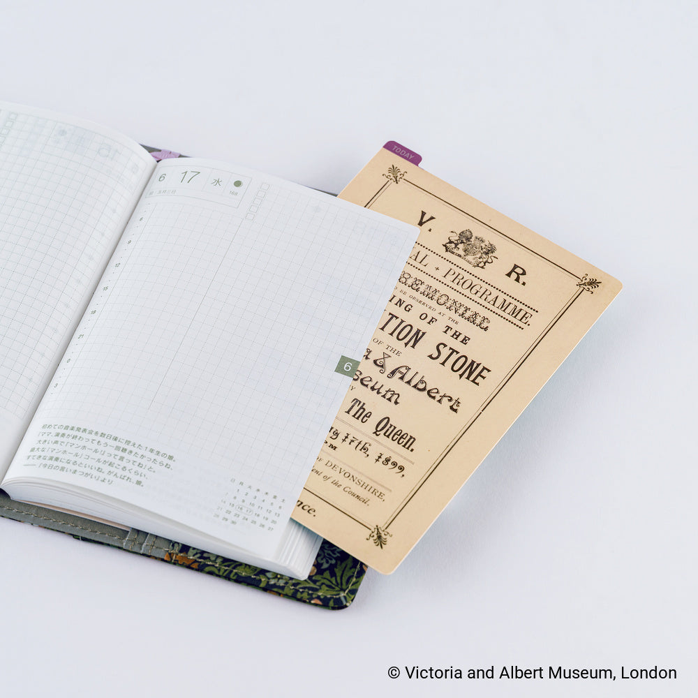 An open planner with grid pages and printed text, layered over a pencil board with the official programme from the Victoria and Albert Museum, London. The shot is styled on a clean white background with a soft shadow.