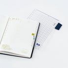 An open planner with a grid page and gold page marker, placed on top of a clear pencil board with a dark blue corner piece and gridded design, on a clean white background.