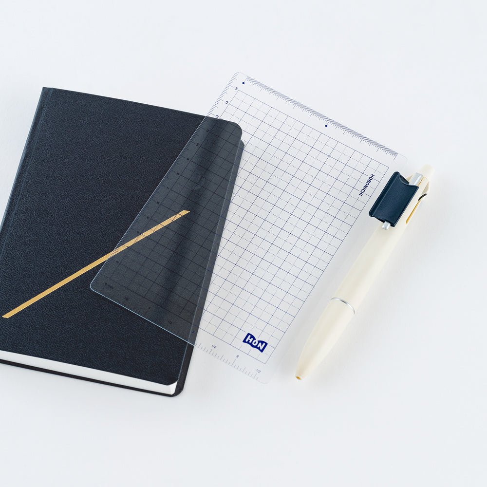 A clear grid pencil board placed on top of a navy HON notebook and next to a cream pen on a white background.