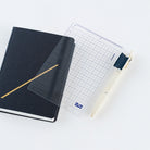 A clear grid pencil board placed on top of a navy HON notebook and next to a cream pen on a white background.