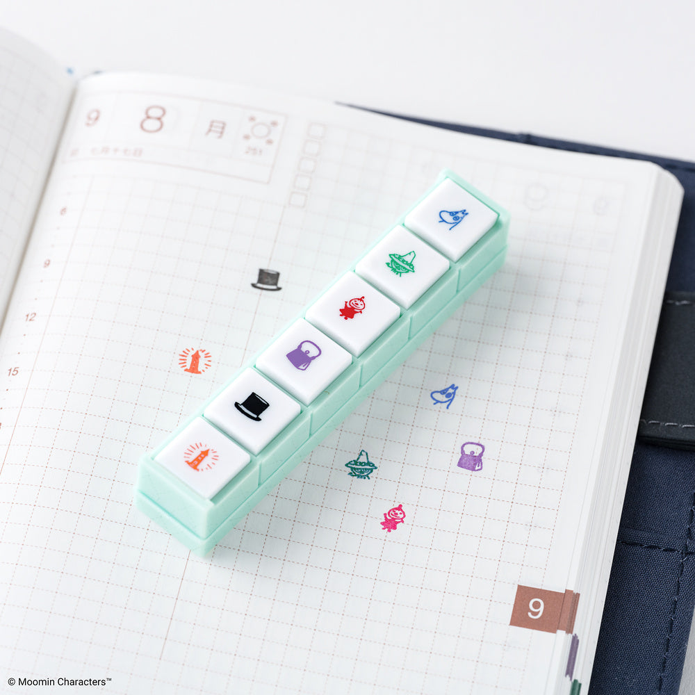 A mint green Pochitto6 push-style button stamp with six Moomin designs against a gridded notebook page