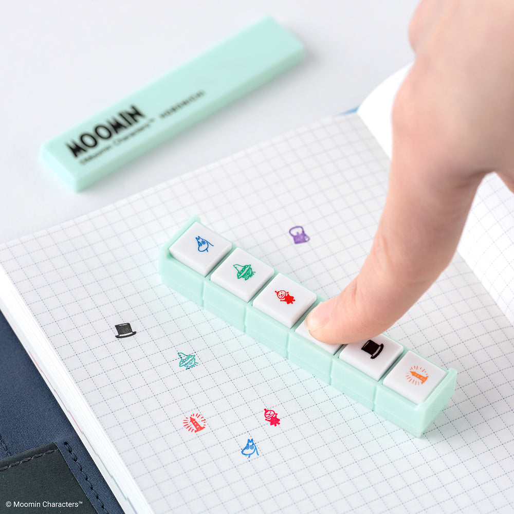 A finger presses a mint green Moomin-themed Pochitto6 stamp on a graph notebook. The pad has six white square buttons with colorful cartoon character icons, and the Moomin brand is visible on the matching mint green case in the background.