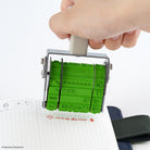 A hand holding a green rubber rotating stamp over a lined notebook, showing the stamped characters and text.