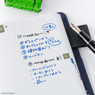 A close-up of a grid-lined planner with black inked headers, handwritten Japanese notes in blue ink, alongside a black fountain pen, a dark navy folio, and a small green stamp at the top edge on a white background.