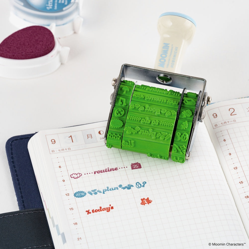 A green rubber stamp ink roller with a metal frame is shown pressed over a planner page. The stamp has cartoon characters embossed in the green rubber.