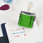 A green rubber stamp ink roller with a metal frame is shown pressed over a planner page. The stamp has cartoon characters embossed in the green rubber.
