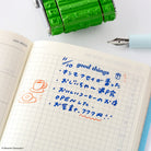 An open grid notebook on a white surface with handwritten Japanese text in blue ink and small orange doodles. A green roller stamp and a light blue fountain pen are placed around it.
