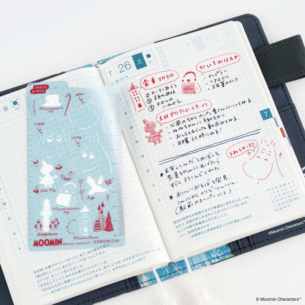 An open planner showing decorated pages with Japanese writing and illustrations inspired by Moomin, including a light blue stencil photographed on a white background.