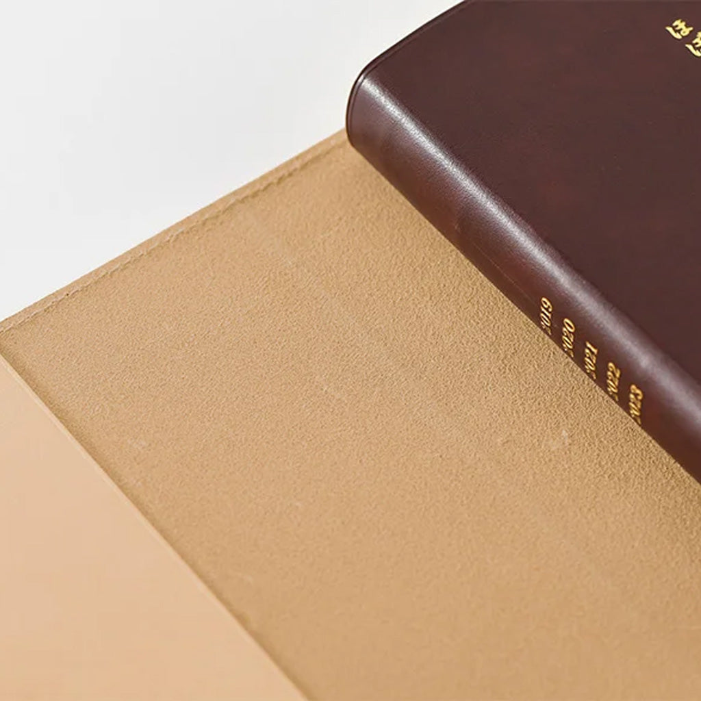 A close-up of a tan textured leather notebook cover with a dark brown book partially placed inside, showing gold text on the spine.