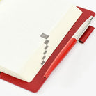 A red leather planner cover holding an open cream-colored grid notebook, with a matching red and silver pen resting on the cover, set against a clean white background.