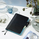 Black and blue notebook with pen on a table setting with plates, glasses, and a vase.