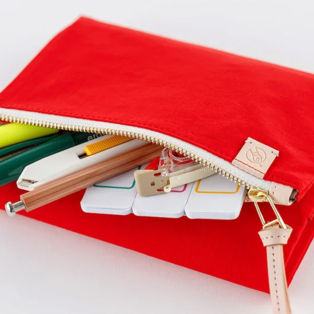 A bright red fabric zip pouch shown open on a white background, with pens, sticky notes, and other stationery items peeking out. A small beige leather tag is attached near the zipper.