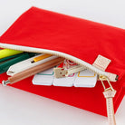 A bright red fabric zip pouch shown open on a white background, with pens, sticky notes, and other stationery items peeking out. A small beige leather tag is attached near the zipper.