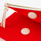 A close-up of a bright red zip pouch with cream-colored leather trim, a cream zipper pull, and gold-tone metal hardware on a white background.