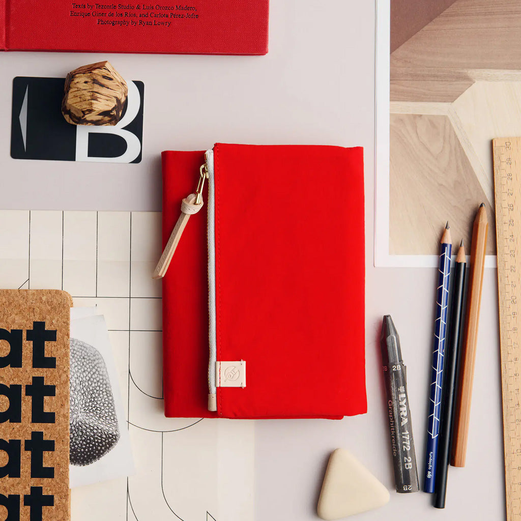 A bright red zippered fabric notebook or planner cover with a small beige leather tag and tassel zipper pull, styled on a white desk flat-lay with pencils, a ruler, a cork board, and other stationery around it.