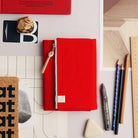 A bright red zippered fabric notebook or planner cover with a small beige leather tag and tassel zipper pull, styled on a white desk flat-lay with pencils, a ruler, a cork board, and other stationery around it.