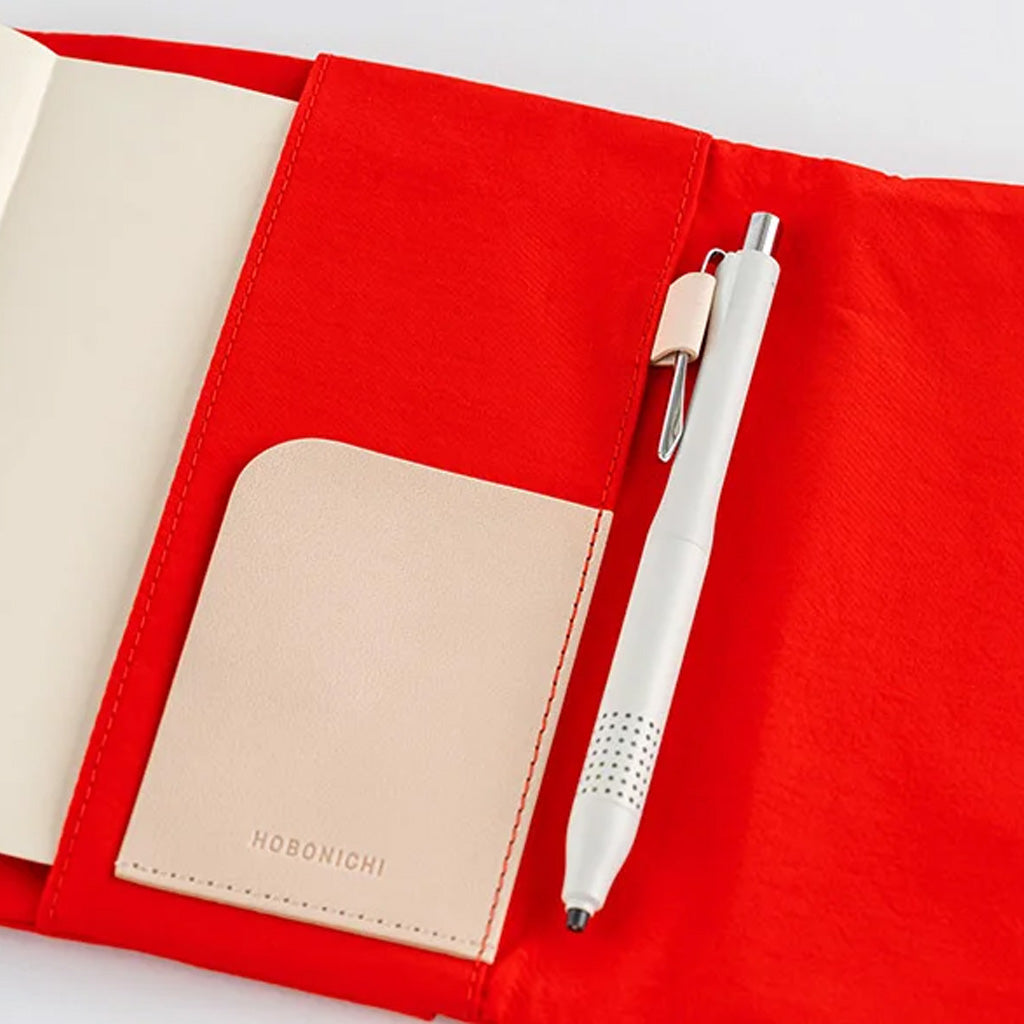 A red fabric notebook cover shown open, with a cream-colored page inside, a beige pocket holding a pen, and a white pen resting on the red surface. The pocket is embossed with the brand name HOBONICHI.