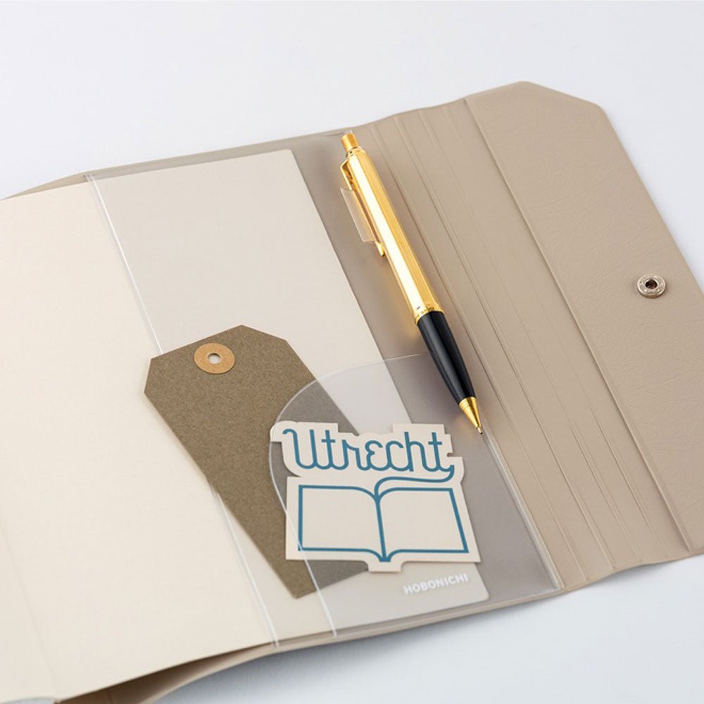 A close up view of an open Hobonichi Techo cover with a pen, tag, and sticker tucked into a clear pvc pocket on a white background.