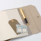 A close up view of an open Hobonichi Techo cover with a pen, tag, and sticker tucked into a clear pvc pocket on a white background.