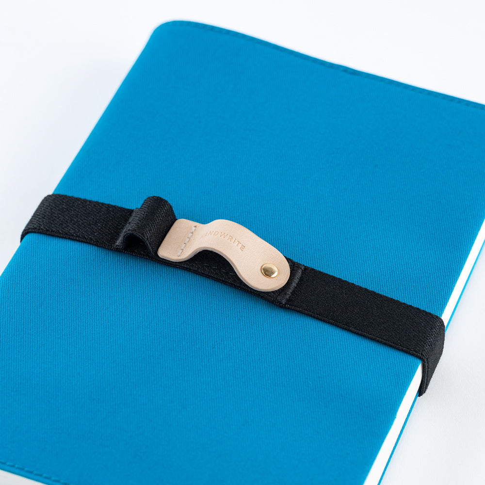 A bright blue planner cover with a black elastic strap and a natural leather pen loop, photographed on a white background.