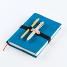 A Hobonichi Techo with a blue cover featuring a removable black elastic band holding two pens on a white background.