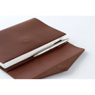 A brown leather Hobonichi Techo cover holding a white planner and a pen in a pen loop, photographed on a white background.