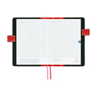 An open planner with white grid pages and a black and interior with red pen closure loops and two ribbon bookmarks hanging below, one red and one pink, on a plain white background.