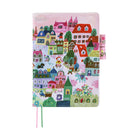 A pink journal cover featuring a whimsical, colorful illustration of a village with houses, trees, and characters. The cover has a stitched edge, a pink pen closure tab on the right, and two ribbon bookmarks in pink and green, shown against a plain white background.