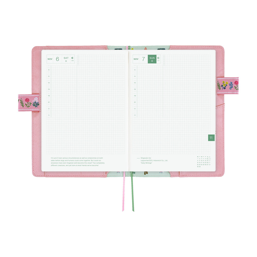 An open planner with a pink cover and white grid pages, shown on a plain white background. The cover has small floral details on the side tabs, and two ribbon bookmarks in pink and green hang from the bottom.