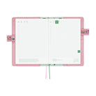 An open planner with a pink cover and white grid pages, shown on a plain white background. The cover has small floral details on the side tabs, and two ribbon bookmarks in pink and green hang from the bottom.