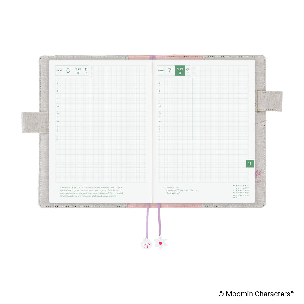An open monthly planner with two grid pages visible, a beige cover, pink and lavender ribbon bookmarks, and decorative charms, shown on a white background.