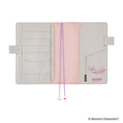 An open Moomin planner cover in a light beige cotton material with a pink fabric accent. It features multiple card slots, pen loops, and  purple ribbon bookmarks with small charms at the ends.