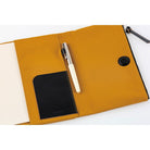 Close-up of the interior of a mustard yellow Hobonichi Tragen cover, showing a black pocket, pen holder with pen, and magnetic closure on a white background.
