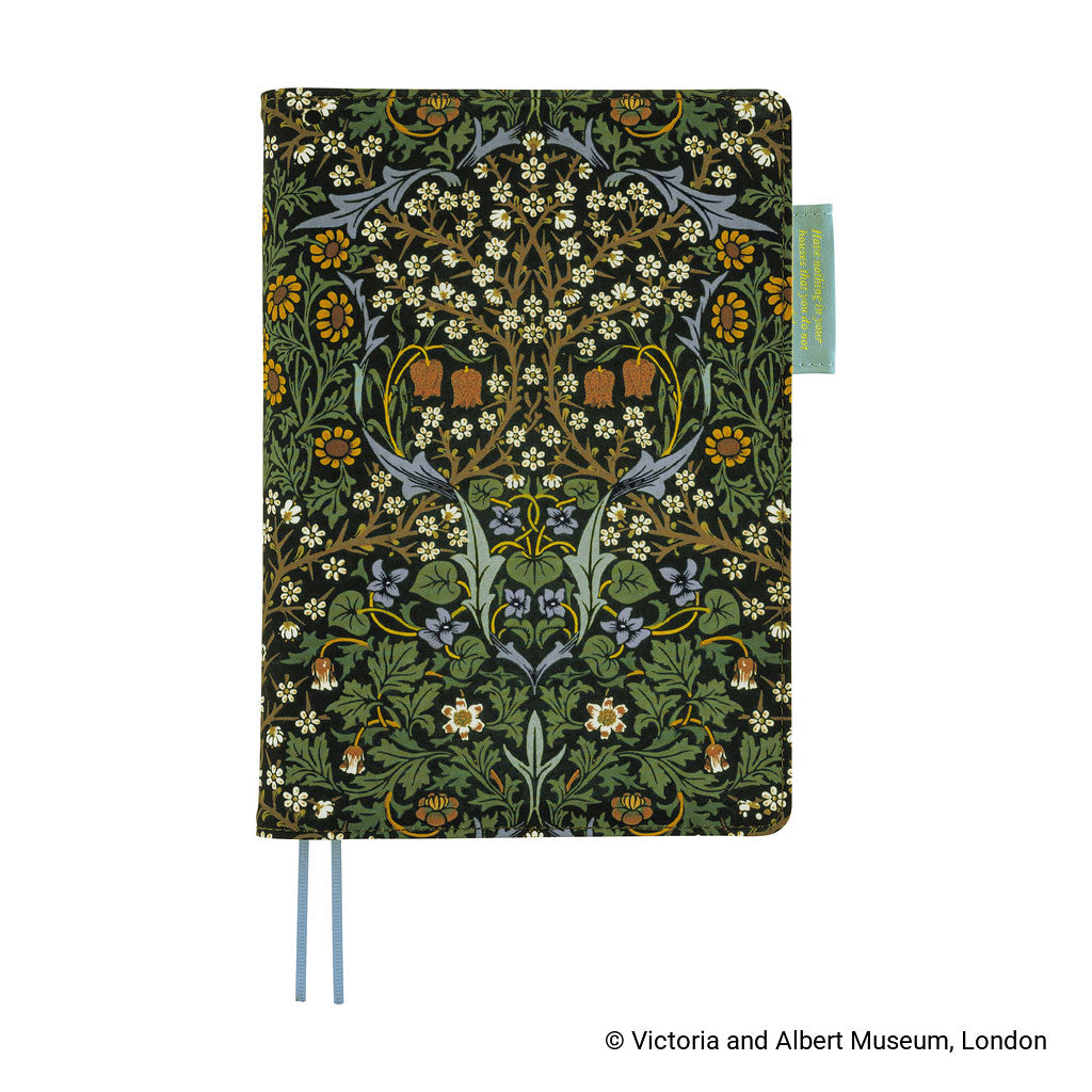 A Hobonichi Techo planner cover featuring a detailed floral and botanical pattern in green, yellow, white, and blue on a black background, with two blue ribbon bookmarks and a small turquoise tag on the right side.