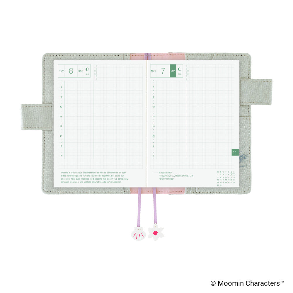 An open monthly planner with two grid pages visible, a pale sage green cover, pink and lavender ribbon bookmarks, and decorative charms, shown on a white background. A Moomin Characters copyright mark appears in the bottom right corner.