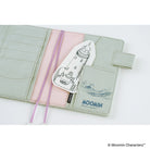 A pale sage green faux-leather card holder wallet with a pink elastic band, featuring a white lighthouse-shaped card with a black line drawing and the blue 'MOOMIN' logo.