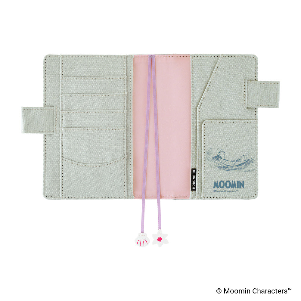 A light sage green Moomin wallet shown open, featuring a pink center pocket, purple drawstring cords with star and shell charms, and a blue Moomin logo on the right pocket.