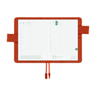 An open grid planner with a reddish-orange cover and strap, showing two white daily pages with green date markers and printed text, photographed on a plain white background.