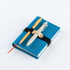 A blue hardcover notebook with a black elastic band closure, holding a wooden pencil and a white pen. The notebook has a red bookmark ribbon peeking out from the left side, photographed on a clean white background.