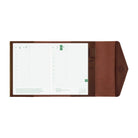 A brown leather planner cover shown open, displaying two white grid planner inserts with green accents on a plain white background.